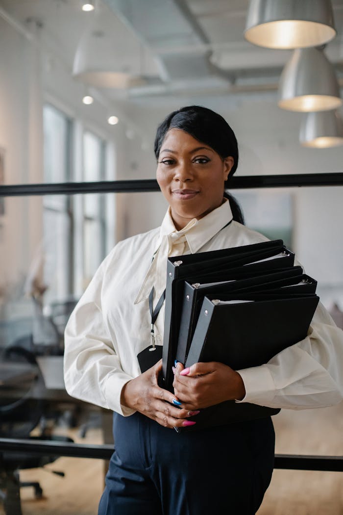 Smiling African American female employee wearing classy outfit standing with paper documents in modern workplace